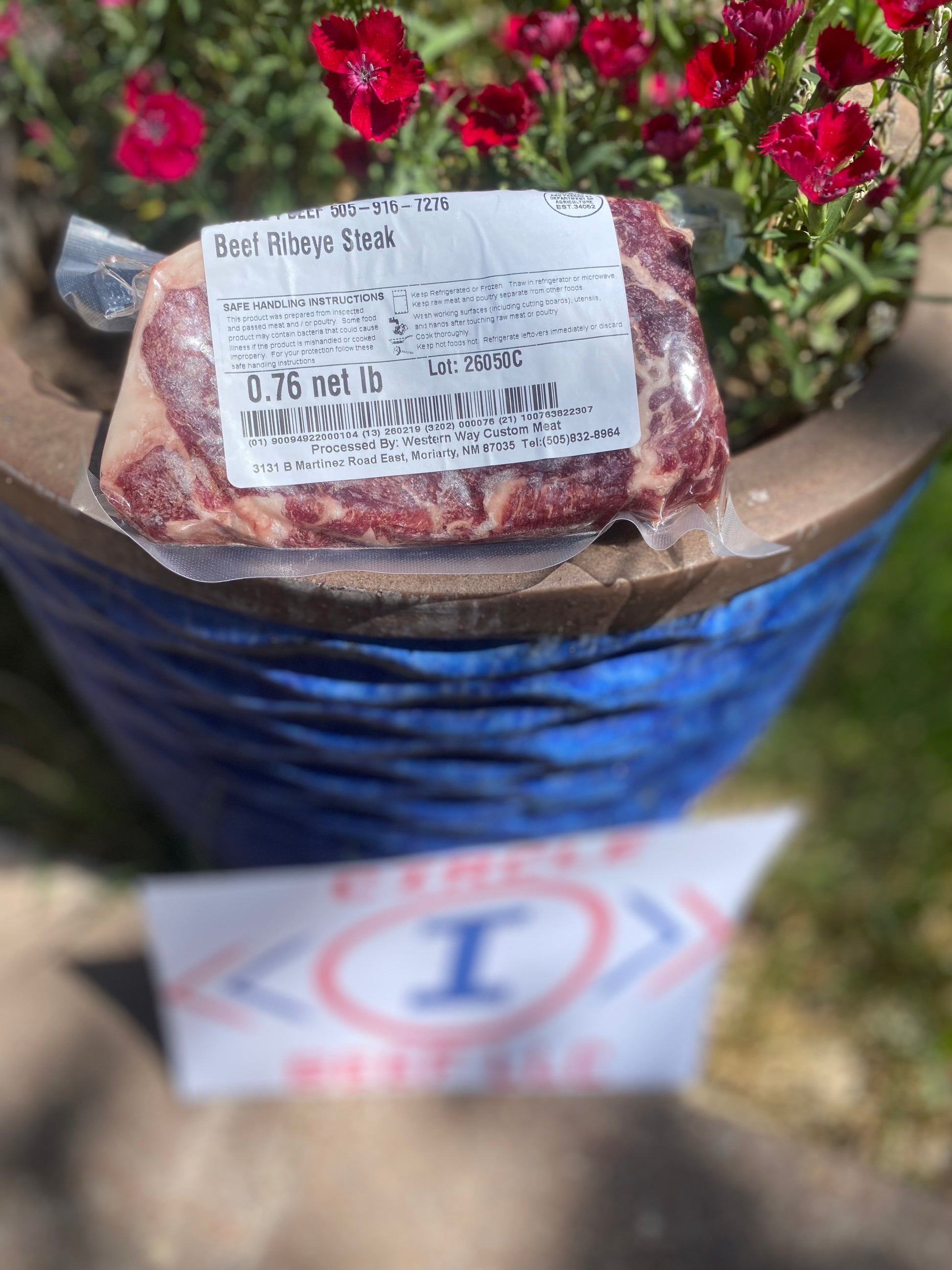 Packaged beef ribeye steak with a label on top of a blue barrel, with flowers in the background.